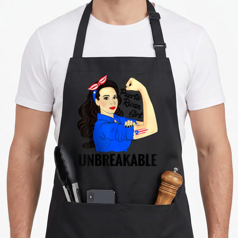 Unbreakable heritage aprons - Cartoon woman flexing arm with Puerto Rican flag tattoo and UNBREAKABLE text — shown on a black cotton canvas apron against a white background