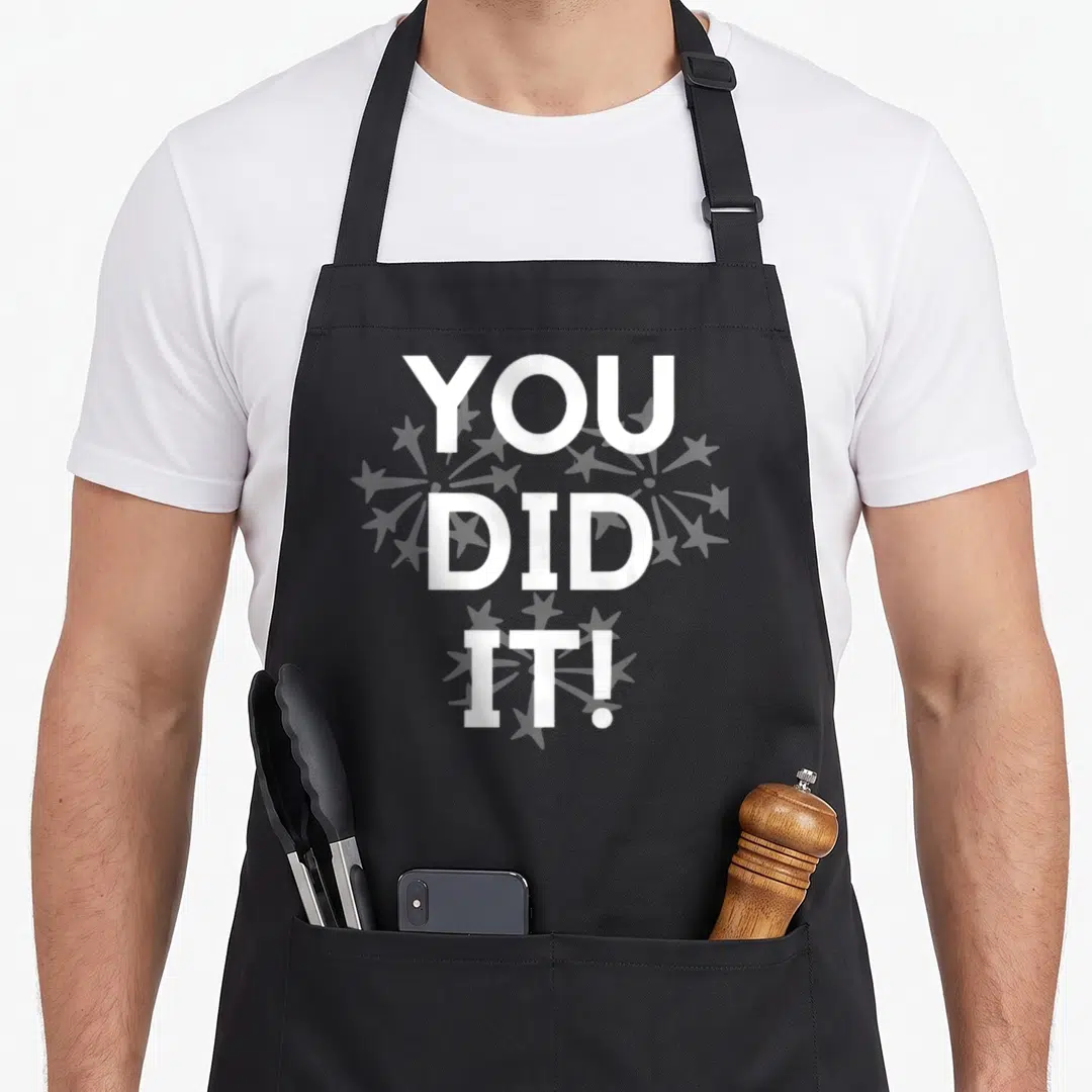 Congratulation congrats apron - White YOU DID IT text with grey dandelion seeds on grey background — shown on a black cotton canvas apron against a white background