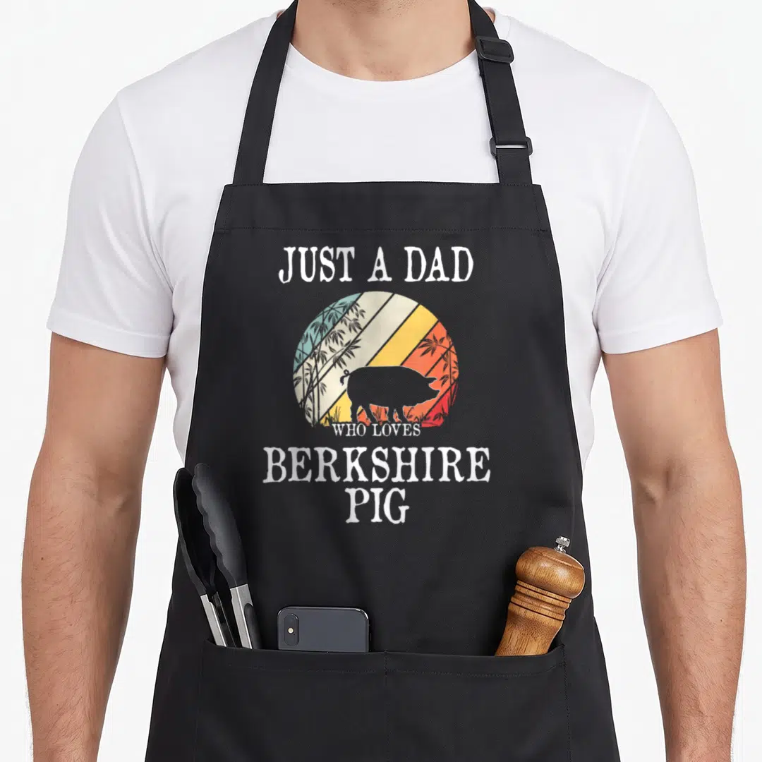 Berkshire pig dad apron - Retro sun with bamboo and pig silhouette flanked by Just A Dad Who Loves Berkshire Pig text — shown on a black cotton canvas apron against a white background