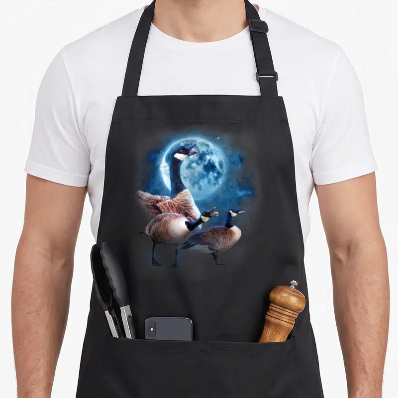 Howling silly apron - Three Canada geese standing under a large blue full moon with a starry sky background — shown on a black cotton canvas apron against a white background
