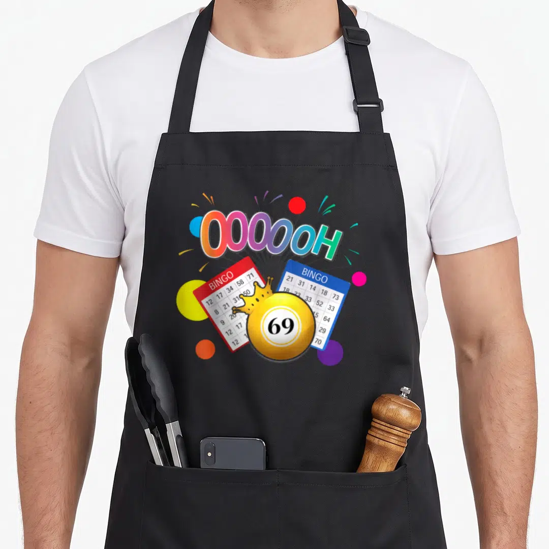 Drag bingo apron - OOOOOH text, yellow 69 ball with crown, red and blue bingo cards, colorful circles — shown on a black cotton canvas apron against a white background