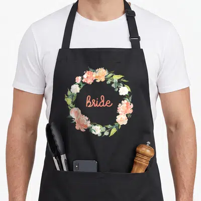 Funny bride wedding day aprons - Coral and white watercolor floral wreath enclosing the word Bride in script lettering — shown on a black cotton canvas apron against a white background