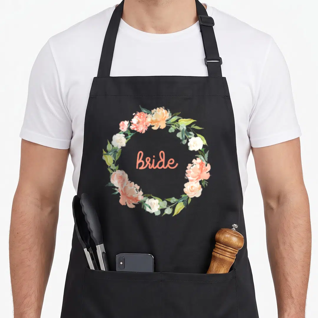 Funny bride wedding day aprons - Coral and white watercolor floral wreath enclosing the word Bride in script lettering — shown on a black cotton canvas apron against a white background