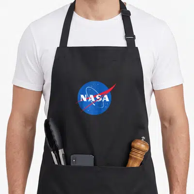Nasa trendy apron - Blue circle with white NASA text red swoosh and orbital ring on grey background — shown on a black cotton canvas apron against a white background