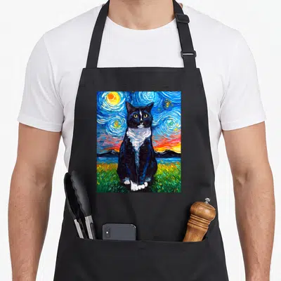 Artwork tuxedo apron - Black and white cat sitting in green grass under a swirling blue yellow starry night sky — shown on a black cotton canvas apron against a white background