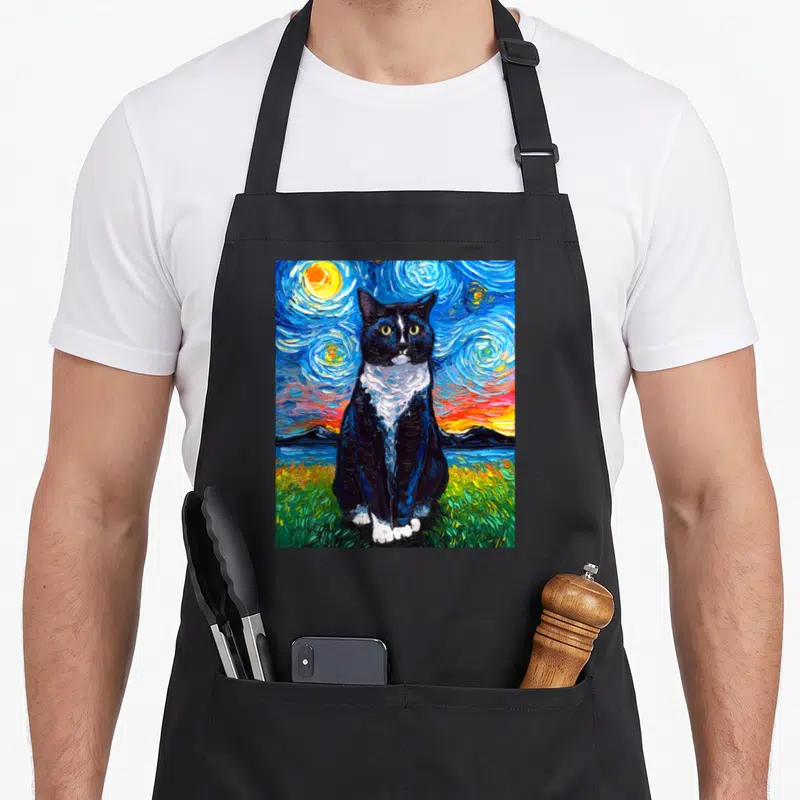 Artwork tuxedo apron - Black and white cat sitting in green grass under a swirling blue yellow starry night sky — shown on a black cotton canvas apron against a white background