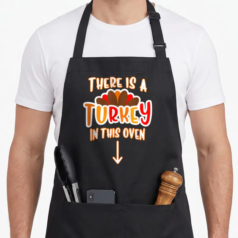 Announcement funny turkey apron - Cartoon turkey head and text THERE IS A TURKEY IN THIS OVEN with downward arrow — shown on a black cotton canvas apron against a white background