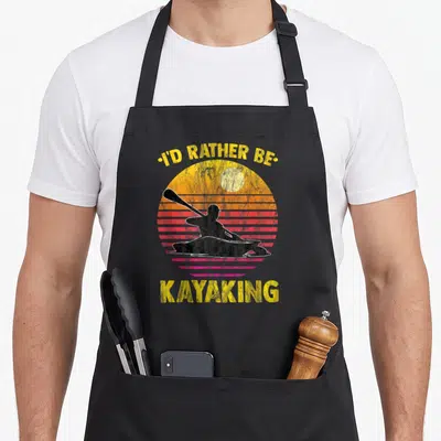 Rather rower apron - I'D RATHER BE KAYAKING text over retro sunset with black kayaker silhouette — shown on a black cotton canvas apron against a white background