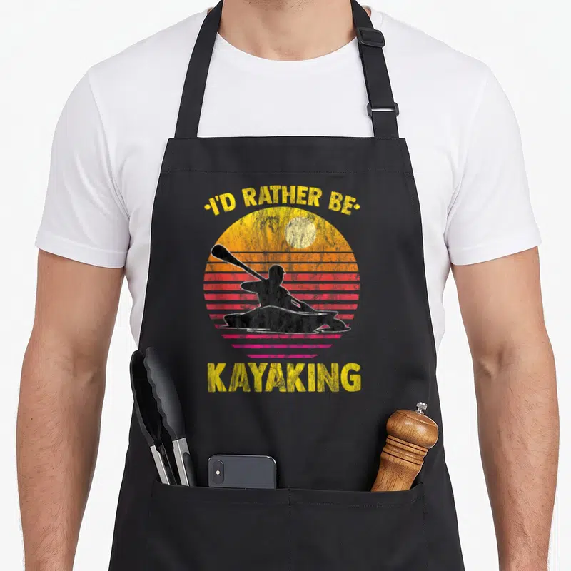 Rather rower apron - I'D RATHER BE KAYAKING text over retro sunset with black kayaker silhouette — shown on a black cotton canvas apron against a white background