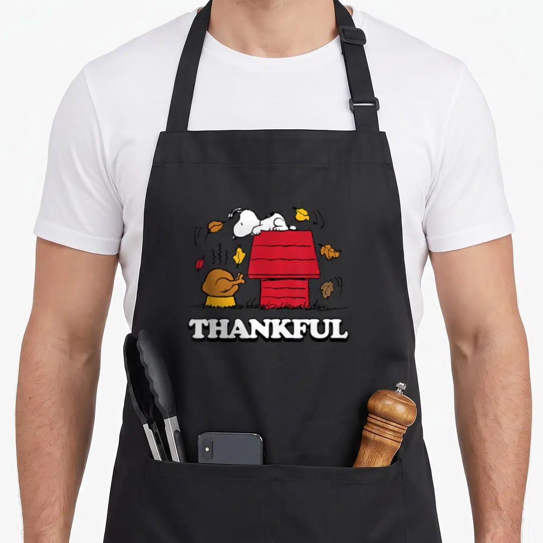 Fall thankful apron - Snoopy on red doghouse with turkey, falling leaves, and THANKFUL text in cartoon style — shown on a black cotton canvas apron against a white background