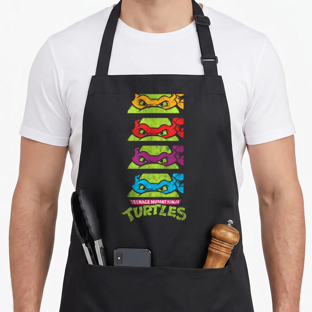 Paneled teenage cooking aprons - Four stacked ninja turtle eyes in orange, red, purple, and blue above TEENAGE MUTANT — shown on a black cotton canvas apron against a white background