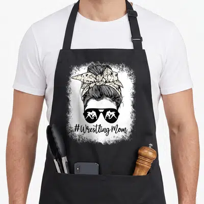 Wrestling messy aprons - #Wrestling Mom text under woman's head with sunglasses reflecting two wrestlers and messy bun — shown on a black cotton canvas apron against a white background