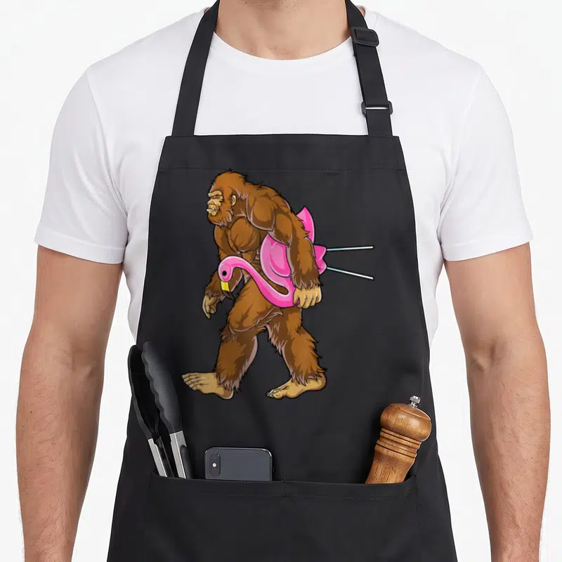 Bigfoot flamingo apron - Brown cartoon bigfoot walking left holding pink flamingo pool toy — shown on a black cotton canvas apron against a white background