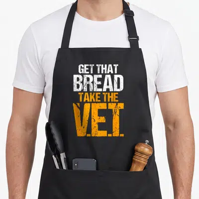 Vet bread apron - Stacked white and orange distressed text reading Get That Bread Take The V.E.I — shown on a black cotton canvas apron against a white background
