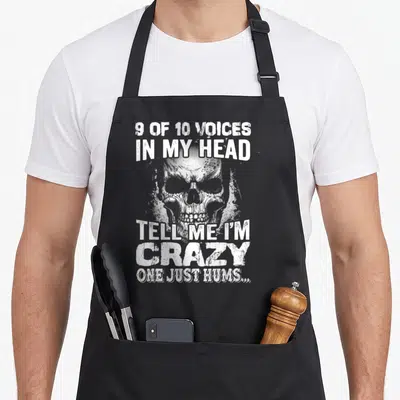 Voice novelty apron - White distressed skull with text 9 of 10 voices in my head tell me i'm crazy one just hums — shown on a black cotton canvas apron against a white background