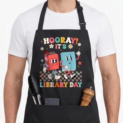 Reading love library apron - Two walking cartoon books with red blue yellow orange pink text and flowers on checkered — shown on a black cotton canvas apron against a white background