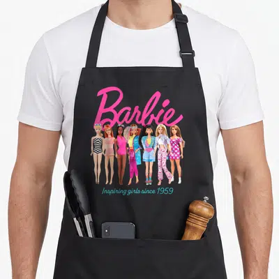 Inspiring doll apron - Pink Barbie script above seven dolls with teal Inspiring girls since 1959 text — shown on a black cotton canvas apron against a white background