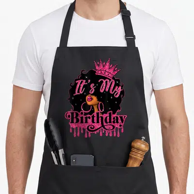 Birthday queen african american aprons - Pink glitter It's My Birthday text over crowned cartoon woman with afro — shown on a black cotton canvas apron against a white background