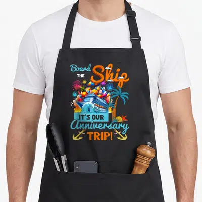Marriage couple apron - Cruise ship surrounded by tropical fruits and fireworks with anniversary trip text — shown on a black cotton canvas apron against a white background