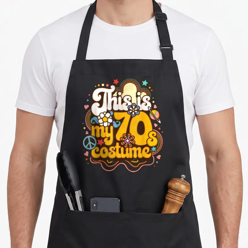 Theme hippie apron - This is my 70s costume text with retro flowers, peace sign, planet, stars, hearts, rainbow border — shown on a black cotton canvas apron against a white background
