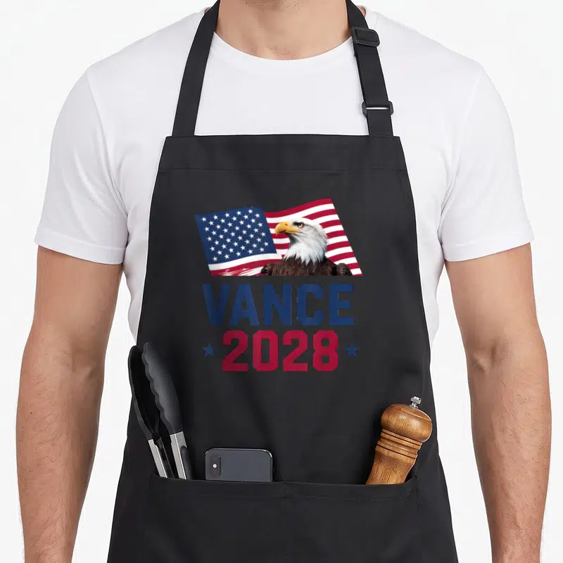 President jd apron - Bald eagle in front of waving USA flag with VANCE 2028 text in blue and red — shown on a black cotton canvas apron against a white background