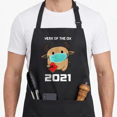 Lunar zodiac aprons - Cartoon brown ox wearing blue mask holding red sign with calligraphy and 2021 text — shown on a black cotton canvas apron against a white background