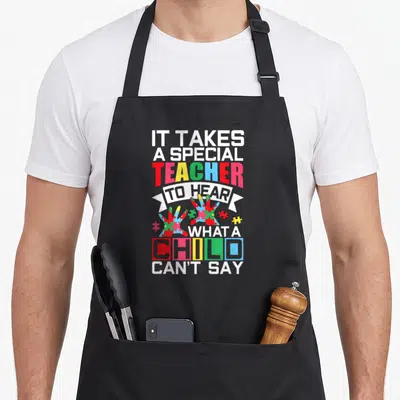 Puzzle teacher art apron - Stacked text IT TAKES A SPECIAL TEACHER TO HEAR WHAT A CHILD CAN'T SAY with puzzle hands — shown on a black cotton canvas apron against a white background