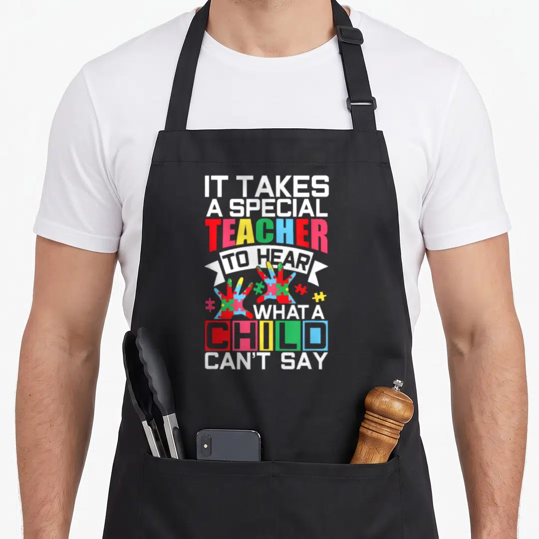 Puzzle teacher art apron - Stacked text IT TAKES A SPECIAL TEACHER TO HEAR WHAT A CHILD CAN'T SAY with puzzle hands — shown on a black cotton canvas apron against a white background
