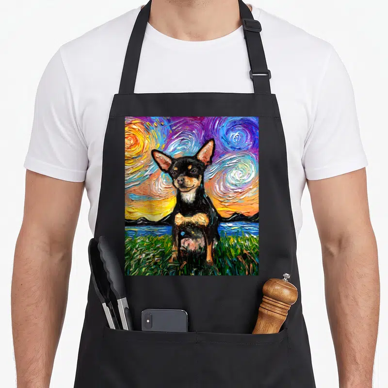 Smooth dog apron - Black and tan chihuahua in colorful swirling starry night landscape with green grass and mountains — shown on a black cotton canvas apron against a white background