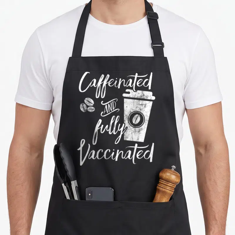 Caffeinated vaccine apron - White distressed text Caffeinated AND fully Vaccinated around coffee cup and beans — shown on a black cotton canvas apron against a white background