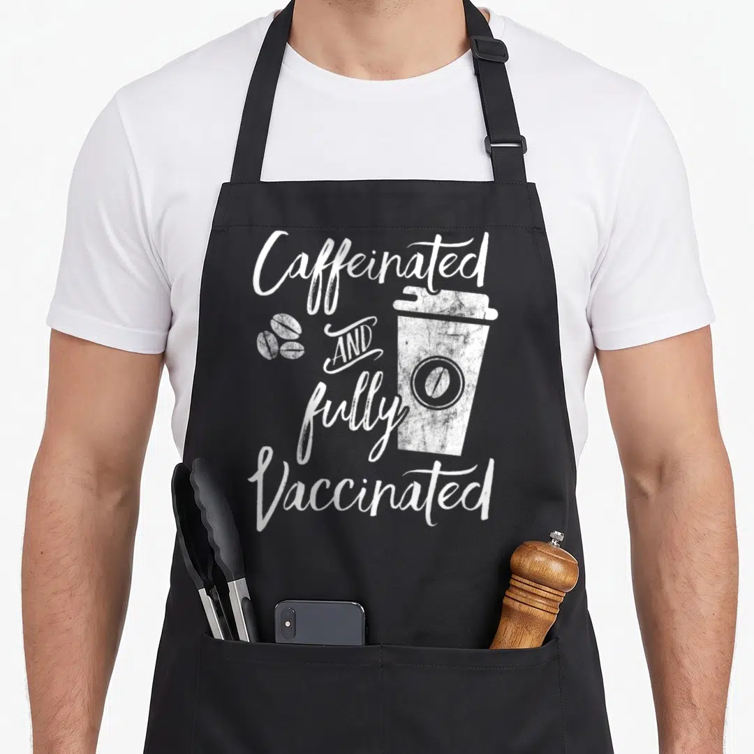Caffeinated vaccine apron - White distressed text Caffeinated AND fully Vaccinated around coffee cup and beans — shown on a black cotton canvas apron against a white background