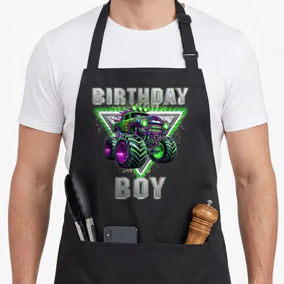 Monster jam truck apron - BIRTHDAY BOY text with cartoon monster truck in silver green purple — shown on a black cotton canvas apron against a white background