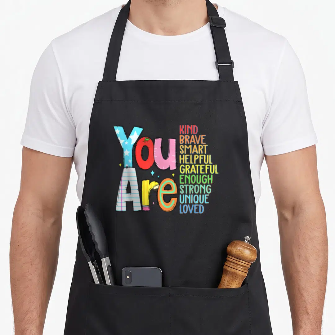Brave teacher apron - You Are text with school objects and colorful adjectives like KIND BRAVE SMART — shown on a black cotton canvas apron against a white background