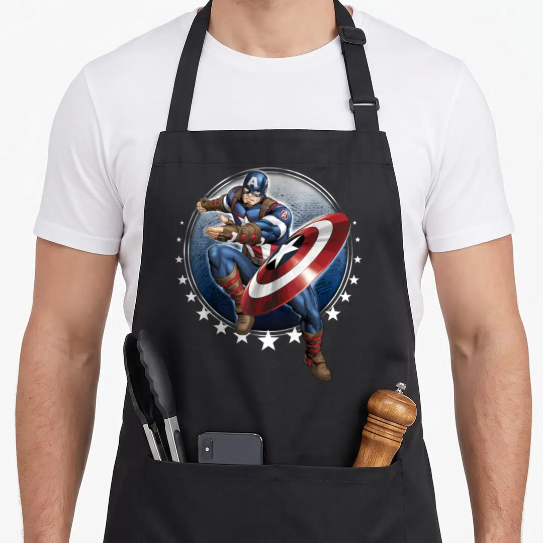 Shield throw apron - Superhero in blue and red holding shield inside silver circle with white stars arching below — shown on a black cotton canvas apron against a white background