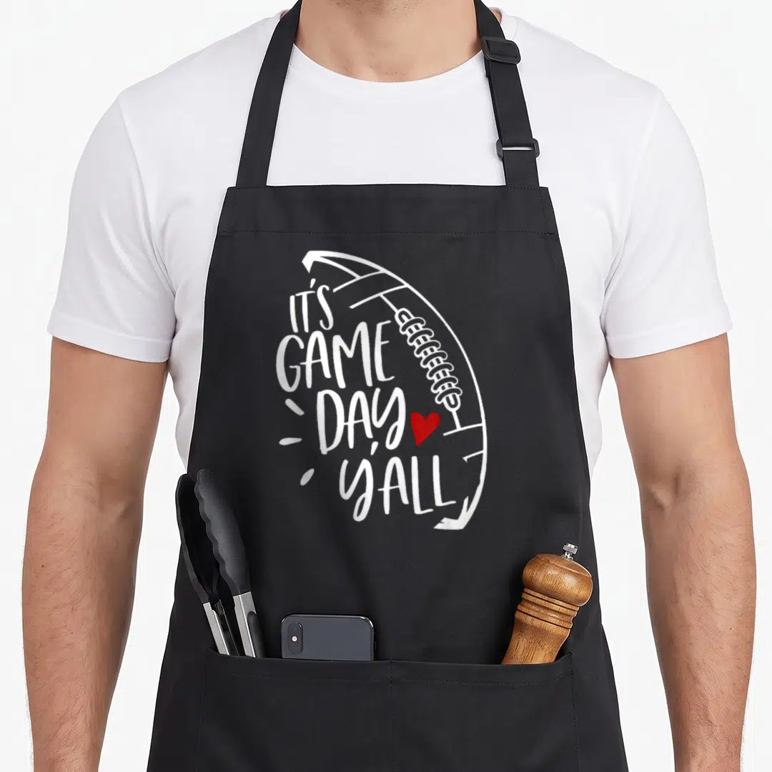 Player game football apron - White text IT'S GAME DAY YALL shaped like a football with a red heart next to DAY — shown on a black cotton canvas apron against a white background