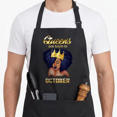 Cute october birthday apron - Cartoon woman with brown skin, blue hair, gold crown, and text Queens are born in OCTOBER — shown on a black cotton canvas apron against a white background