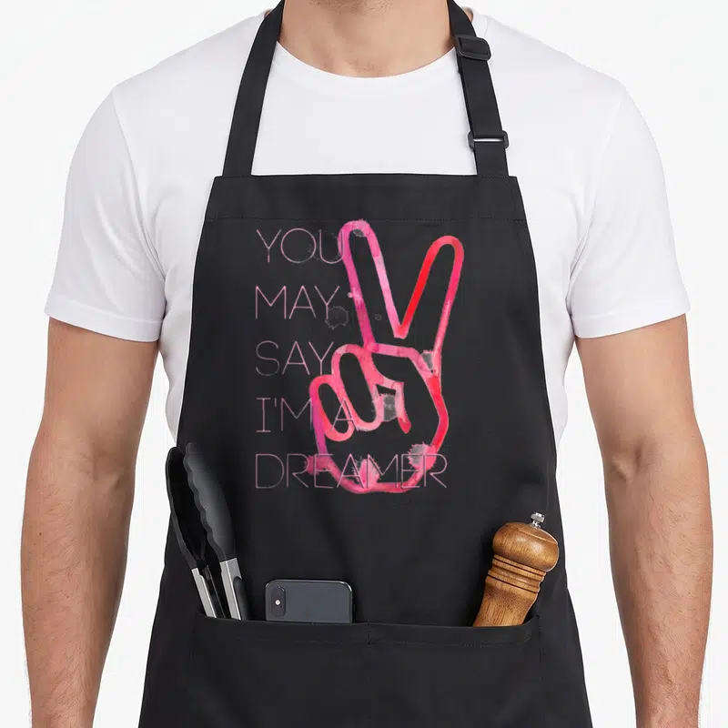 John dreamer apron - Stacked pink text next to a red watercolor peace sign hand with a signature below — shown on a black cotton canvas apron against a white background