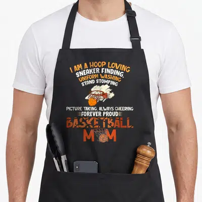 Forever support your apron - Stacked text reading I am a hoop loving basketball mom with two hoop graphics — shown on a black cotton canvas apron against a white background