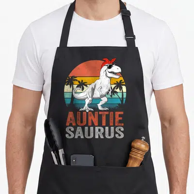 Auntiesaurus rex dinosaur apron - Retro Tyrannosaurus Rex with sunglasses and red bandana standing before striped sun — shown on a black cotton canvas apron against a white background
