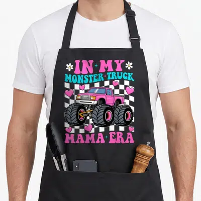 Groovy monster truck apron - Pink monster truck over checkered pattern with In My Monster Truck Mama Era text and hearts — shown on a black cotton canvas apron against a white background