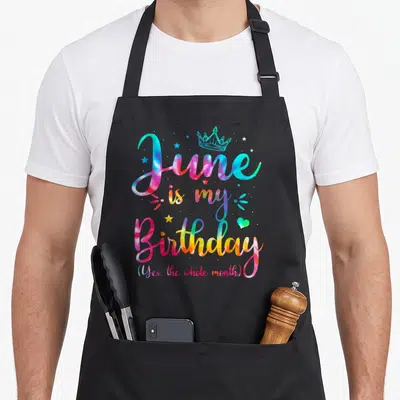 June whole birthday apron - Tie-dye text reading June is my Birthday with crown and stars — shown on a black cotton canvas apron against a white background