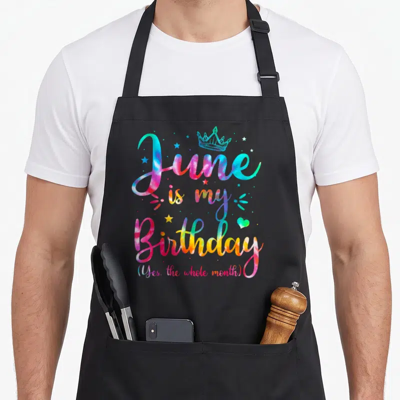 June whole birthday apron - Tie-dye text reading June is my Birthday with crown and stars — shown on a black cotton canvas apron against a white background