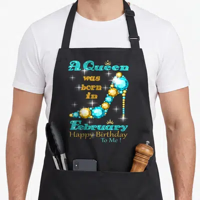 February baking queen apron - A Queen was born in February Happy Birthday To Me text with diamond high heel shoe — shown on a black cotton canvas apron against a white background