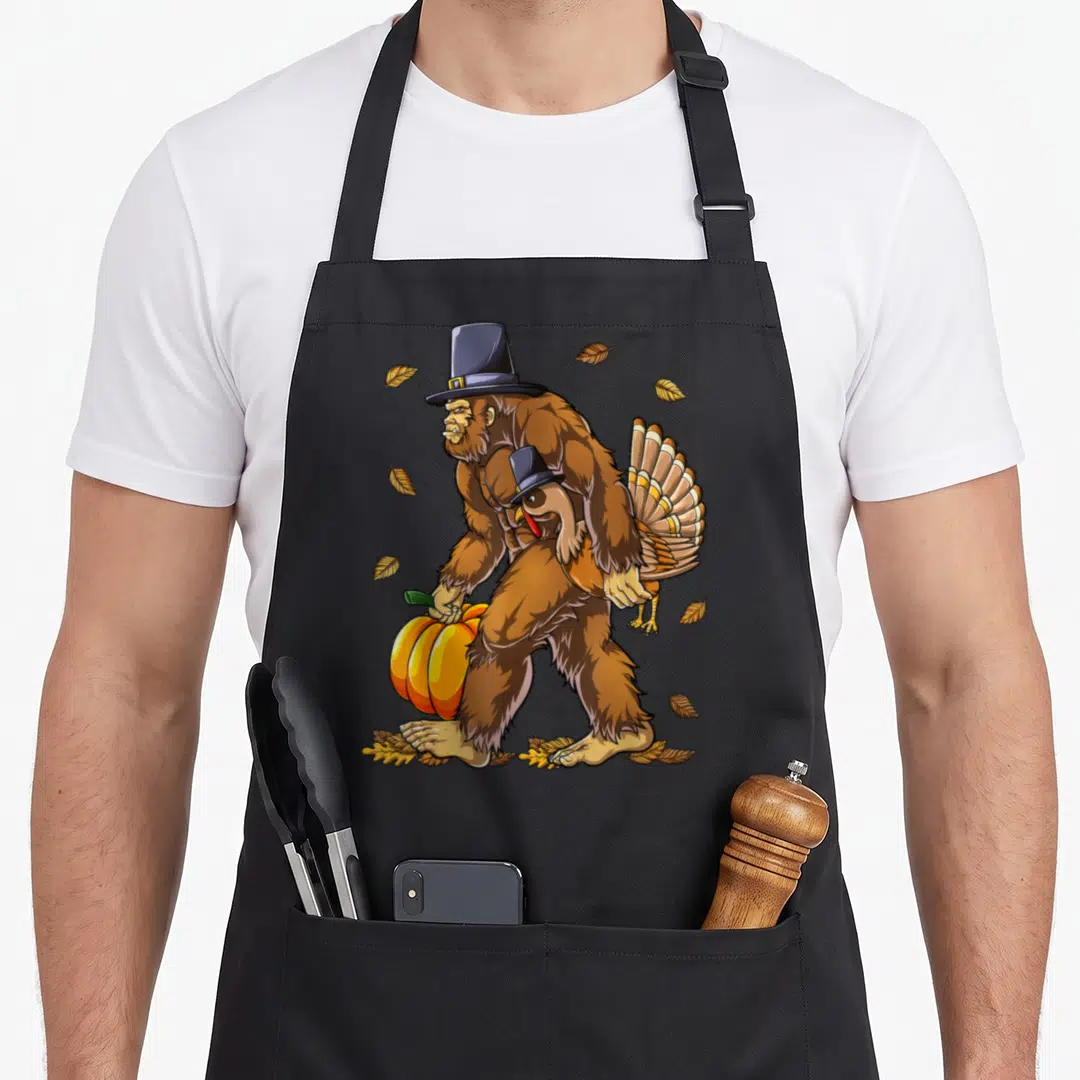 Bigfoot thanksgiving day aprons - Walking Bigfoot in pilgrim hat carrying pumpkin and turkey with scattered leaves — shown on a black cotton canvas apron against a white background