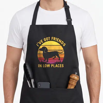 Friend dachshund apron - Grey dog silhouette over yellow orange red sunset stripes with pun text — shown on a black cotton canvas apron against a white background