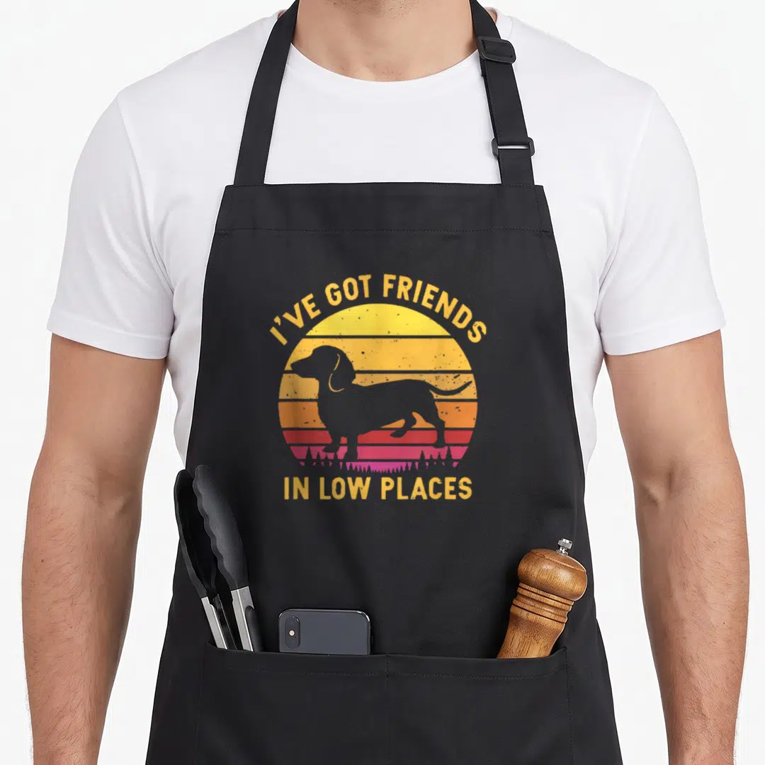 Friend dachshund apron - Grey dog silhouette over yellow orange red sunset stripes with pun text — shown on a black cotton canvas apron against a white background