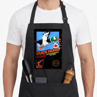 Nes duck apron - Pixelated duck, DUCK HUNT text, light gun icon, gold seal on black background — shown on a black cotton canvas apron against a white background