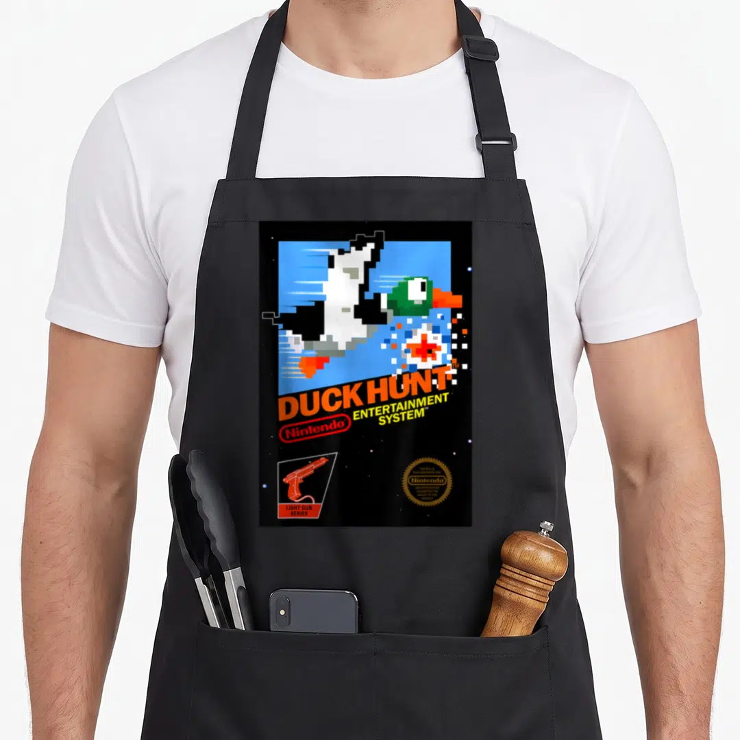 Nes duck apron - Pixelated duck, DUCK HUNT text, light gun icon, gold seal on black background — shown on a black cotton canvas apron against a white background
