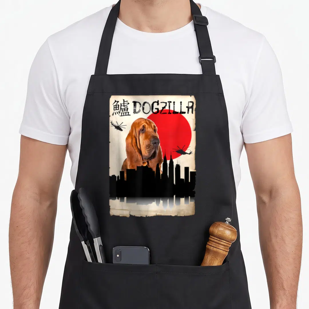 Dogzilla dog apron - Bloodhound dog in front of red sun with DOGZILLA text and two helicopters over black skyline — shown on a black cotton canvas apron against a white background