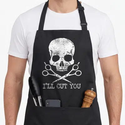 Hairdresser hairstylist apron - White distressed skull and crossed scissors with text I'll Cut You — shown on a black cotton canvas apron against a white background